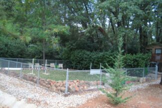 The final area surrounded by a cyclone fenceline, newly installed TuFFGrass artificial turf, elevated with a stacked moss rock edge to level. Ready for agility training for two standard poodles and their owner.